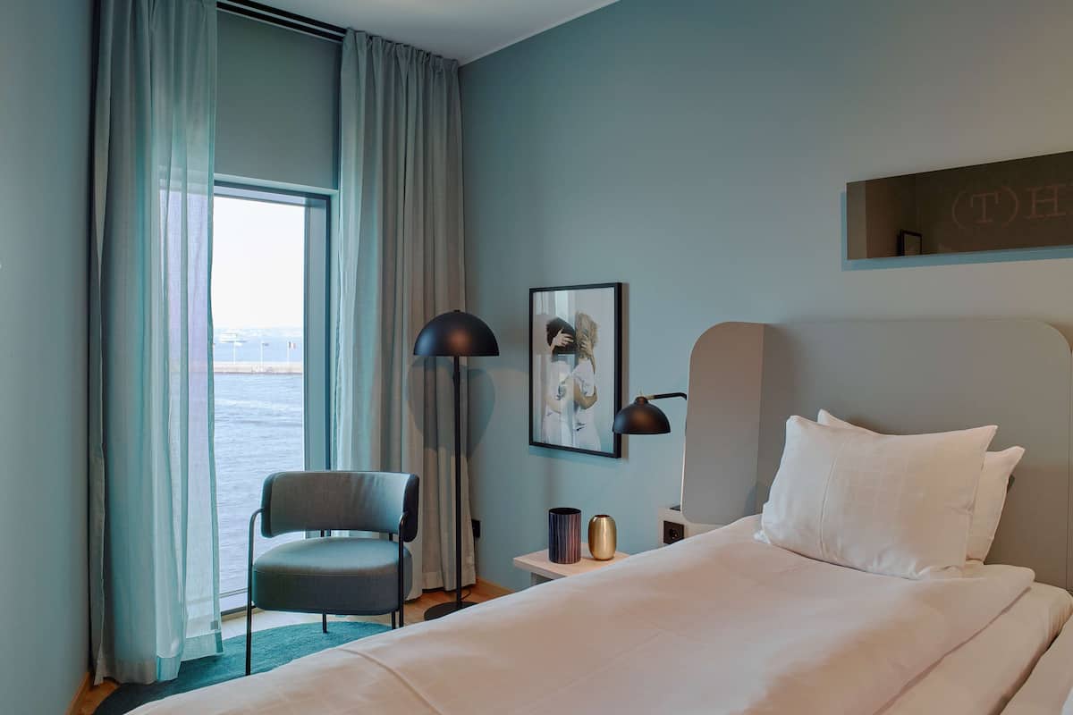 Una habitación de hotel con cama, una silla, un escritorio, una lámpara, un cuadro enmarcado y una ventana con cortinas.