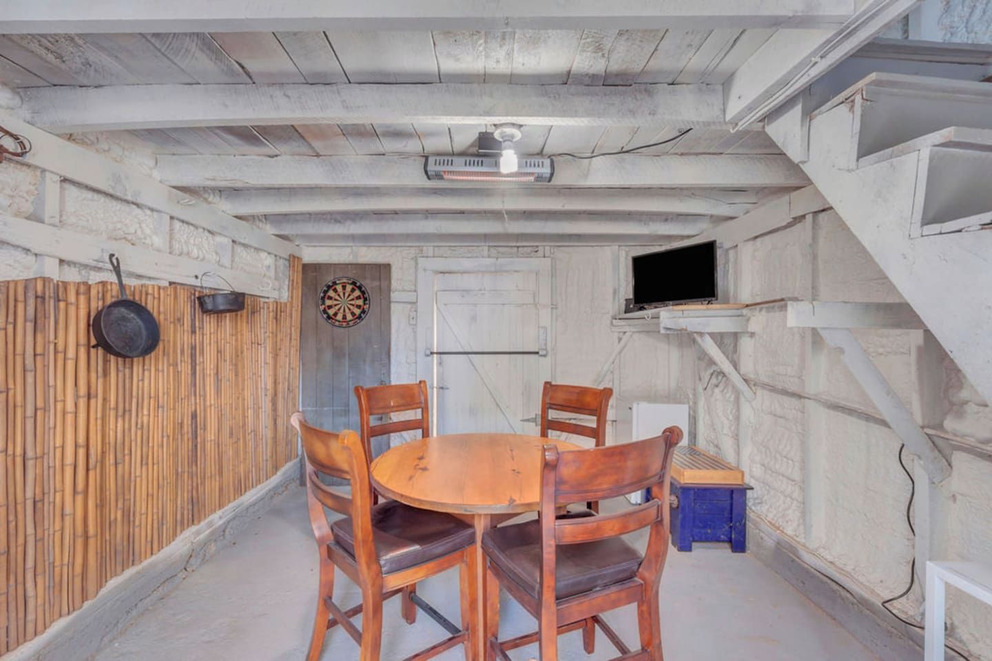 A room with a wooden table and chairs, a dartboard, a television on a shelf, and a door.