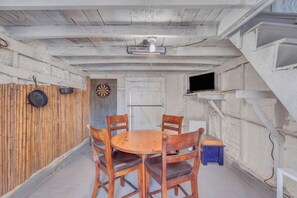 Une pièce avec une table et des chaises en bois, un jeu de fléchettes, un téléviseur posé sur une étagère et une porte.