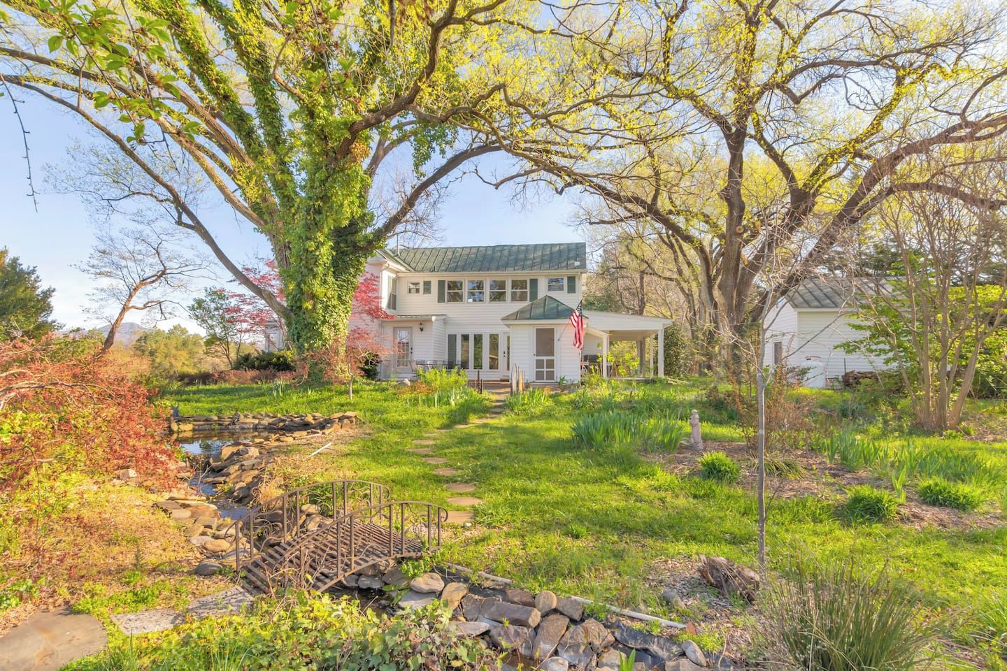 A two-story house with a covered porch, a stone pathway, and a small stream.