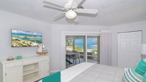 Un dormitorio con cama, ventilador de techo, televisión y vista a la playa a través de una puerta corrediza.