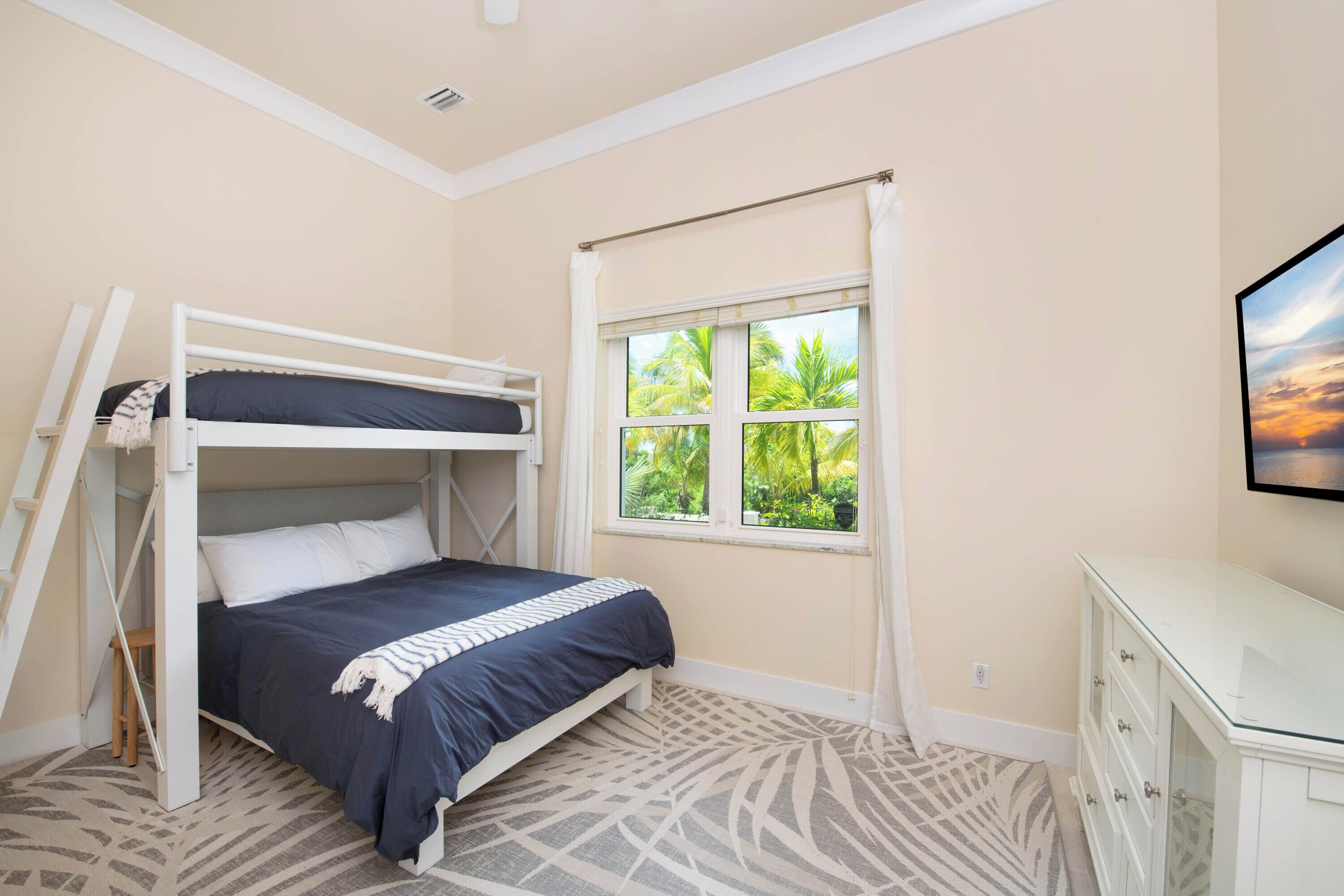 A bedroom with a bunk bed, a window with a view of greenery, a dresser, and a patterned carpet.