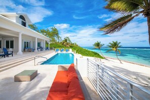A pool area with a view of the ocean, a white building, and a red sofa. - Ocean Kai: Luxe Oasis w/Oceanfront Pool, Sandy Beachfront, & Veranda Lounge (Rum Point)
