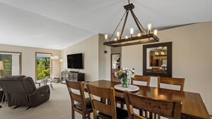 Un comedor con una mesa y sillas de madera, una araña, un televisor de pantalla plana y vistas al exterior con vegetación.