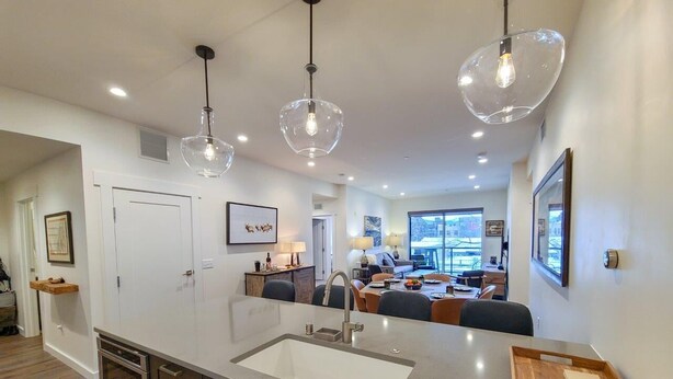 A modern kitchen with a large island, stainless steel appliances, pendant lighting, and a view into the living area.