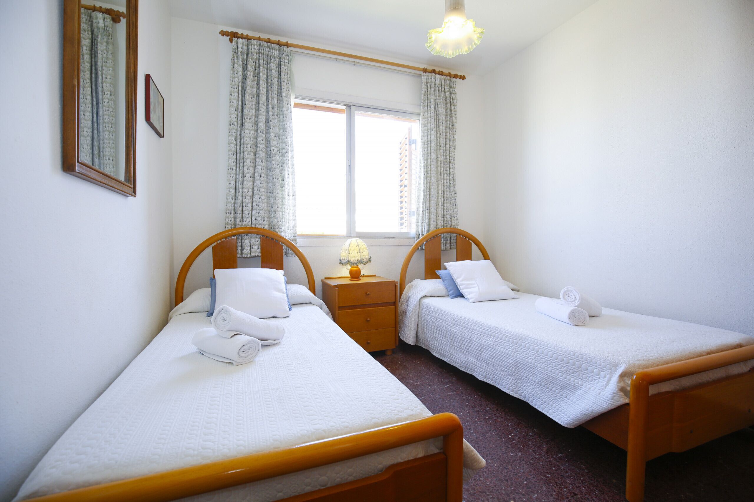 Une chambre avec deux lits simples, une table de chevet avec une lampe, une fenêtre avec des rideaux et un miroir au mur.