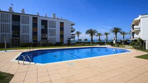 Uma piscina com deck ladrilhado, cercada por palmeiras e um edifício com varandas.