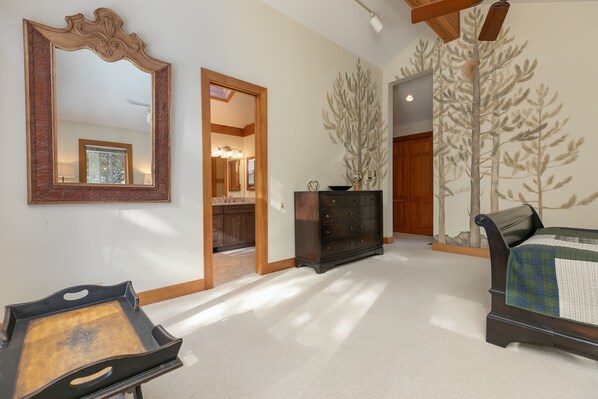 A spacious room with a large mirror, a dresser, and a bed. There is a door leading to another room and a wall with a tree mural.