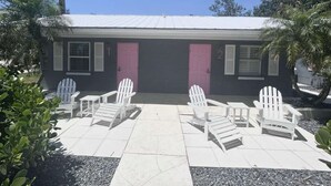 A two-story building with numbered doors, a tiled patio with white Adirondack chairs, and surrounding greenery. - Newly Renovated Charlotte Harbor - Sunset Inn #5 (Port Charlotte)