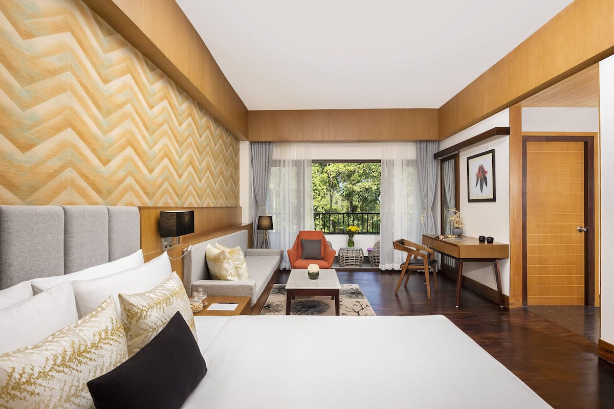 A modern hotel room with a large bed, a sofa, a dining table, and a chair. There is a window with a view of greenery outside.