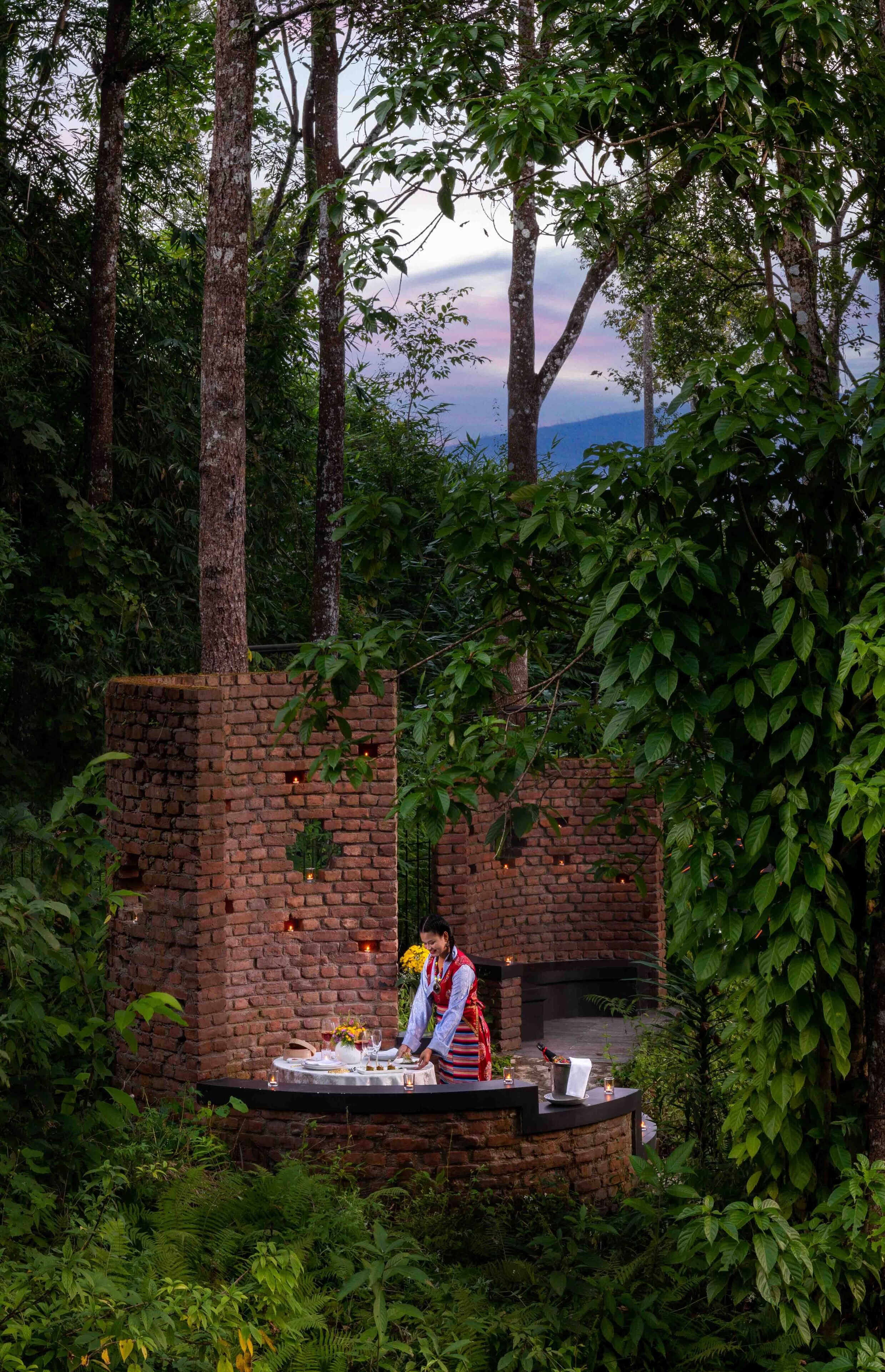 Un espacio comedor al aire libre rústico con una estructura de ladrillo, rodeado de abundante vegetación y árboles.