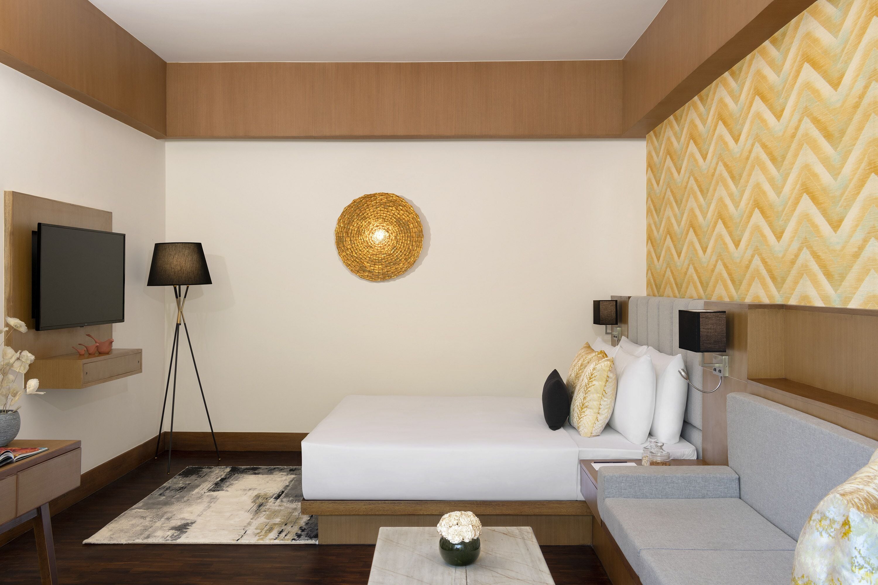 A modern hotel room with a bed, a sofa, a TV, a lamp, and a decorative wall piece.