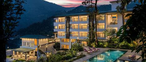 A hotel with a swimming pool, surrounded by lush greenery and a mountainous backdrop.