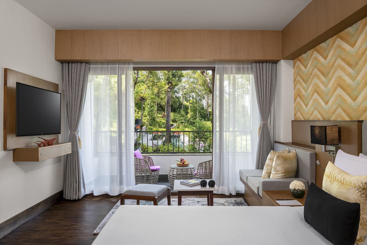 A hotel room with a large TV, a sofa, a dining table, and a balcony with a view of trees.