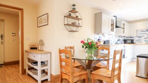 A dining area with a glass table surrounded by wooden chairs, a white cabinet with shelves, and a decorative wall shelf. - Little Gem (Uttoxeter)