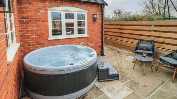 A hot tub next to a brick house with a window, wooden deck, and outdoor furniture. - Little Gem (Uttoxeter)