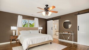 A bedroom with a bed, a ceiling fan, a nightstand, a bedside table, a window with curtains, a mirror, and a door.