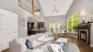 A spacious living room with a high ceiling, a fireplace, and a dining area with natural light from large windows.