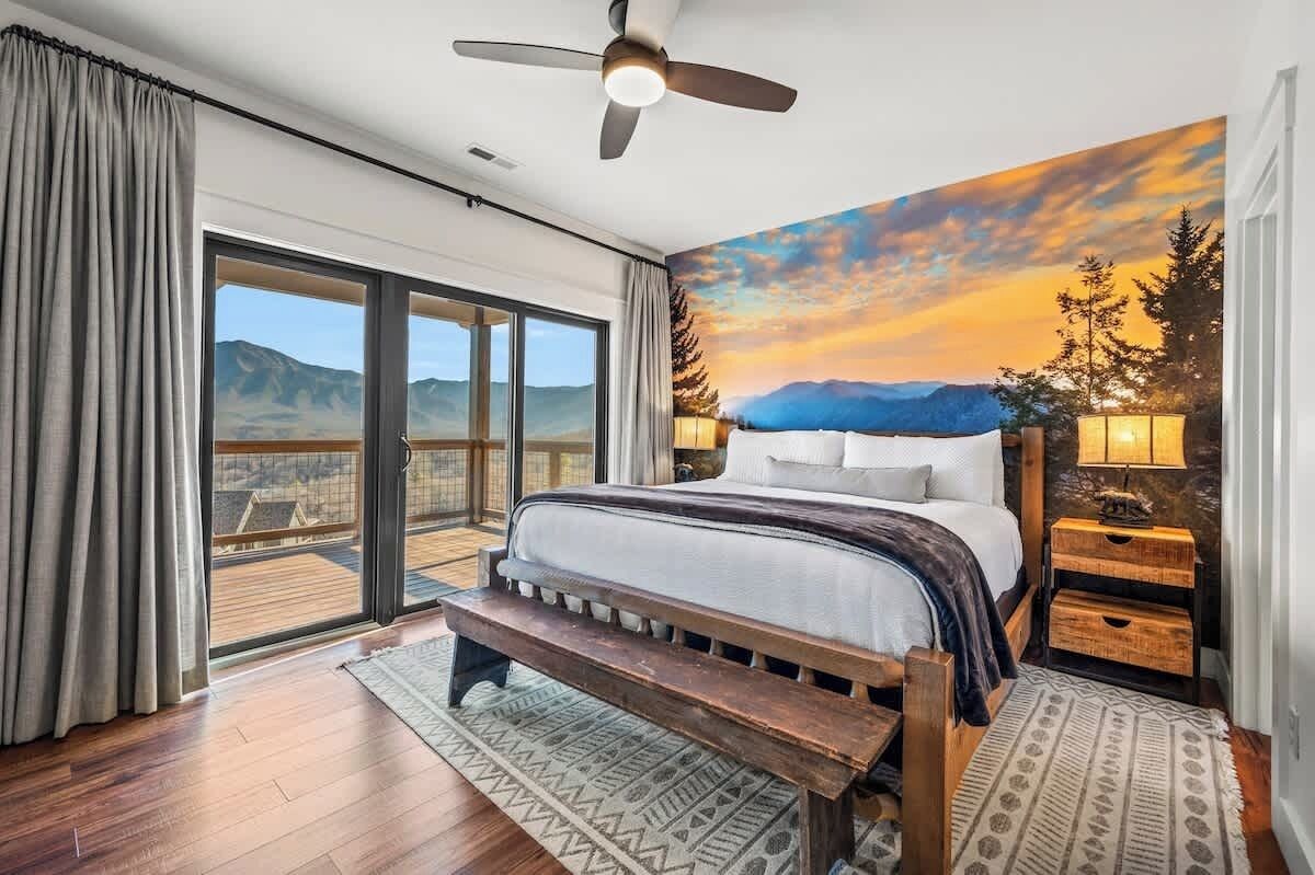 Un dormitorio con una cama grande, un ventilador de techo, una mesita de noche con lámpara y vistas a las montañas a través de un ventanal corredero.