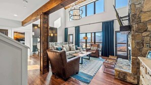 A spacious living area with a stone fireplace, large windows, and a high ceiling with exposed beams.