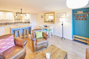 Une cuisine moderne avec des armoires blanches, un comptoir en bois et un luminaire suspendu. On y trouve un coin repas avec une table et des chaises, ainsi qu’un mur bleu orné d’une étagère en bois.
