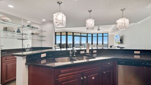 Eine moderne Küche mit dunklen Holzschränken, einer Granit-Arbeitsplatte und einer großen Kochinsel mit Spüle. Es gibt Pendelleuchten mit einem marmorähnlichen Design, und ein großes Fenster mit Blick aufs Meer.