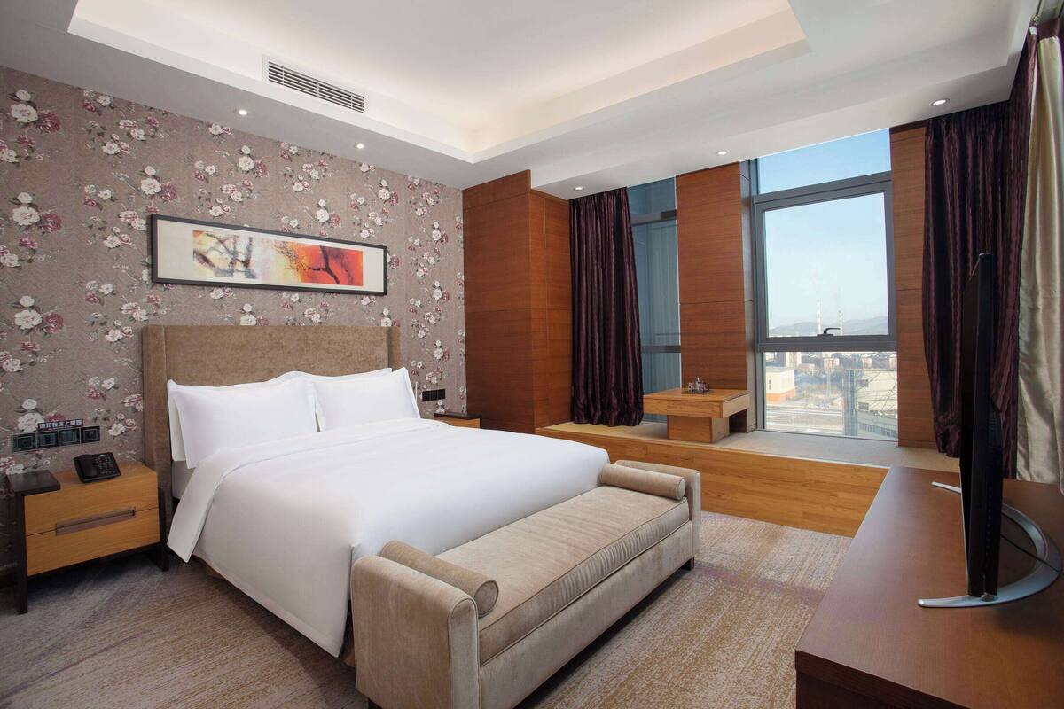 A modern hotel room with a large bed, a bedside table, a flat-screen TV, a window with a city view, and a floral-patterned wall.
