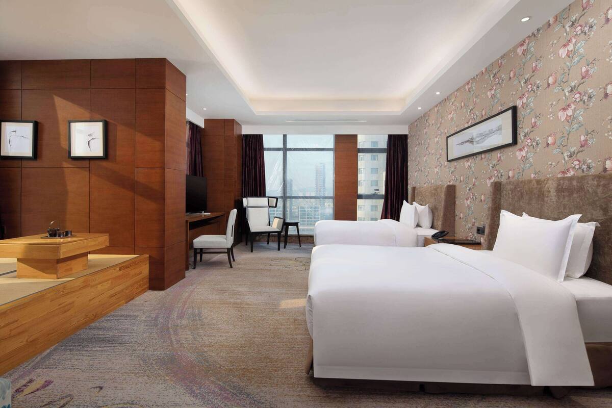 A hotel room with two beds, a desk, a chair, a TV, and a large window with a city view.