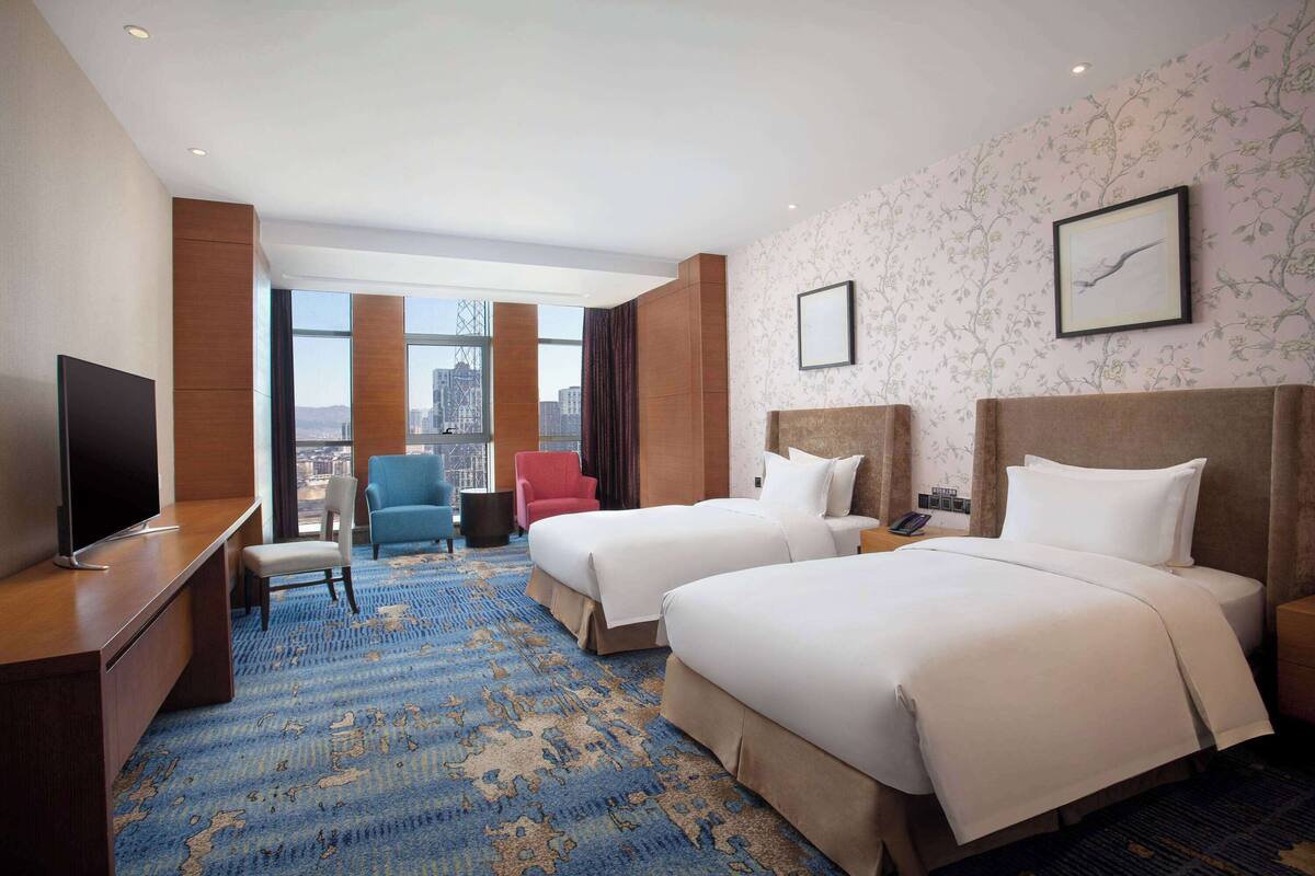 A hotel room with two beds, a desk with a TV, a blue carpet, and a city view.