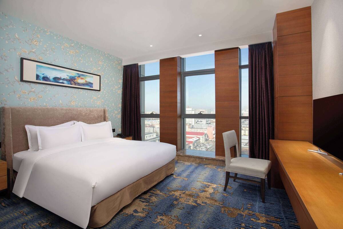 A hotel room with a large bed, a desk, and a chair. There is a large window with curtains and a city view.