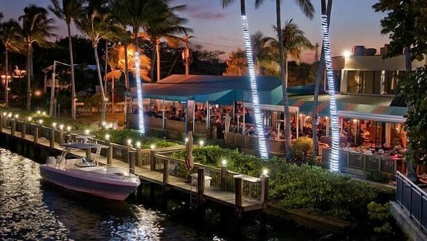 A waterfront restaurant with a dock, palm trees, and a boat.