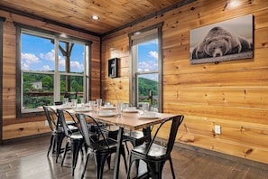 Um interior de madeira com uma mesa de jantar posta para seis pessoas, janelas amplas e um quadro de urso na parede.