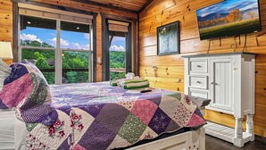 A wooden cabin bedroom with a bed, a TV, and a view of trees through the window.