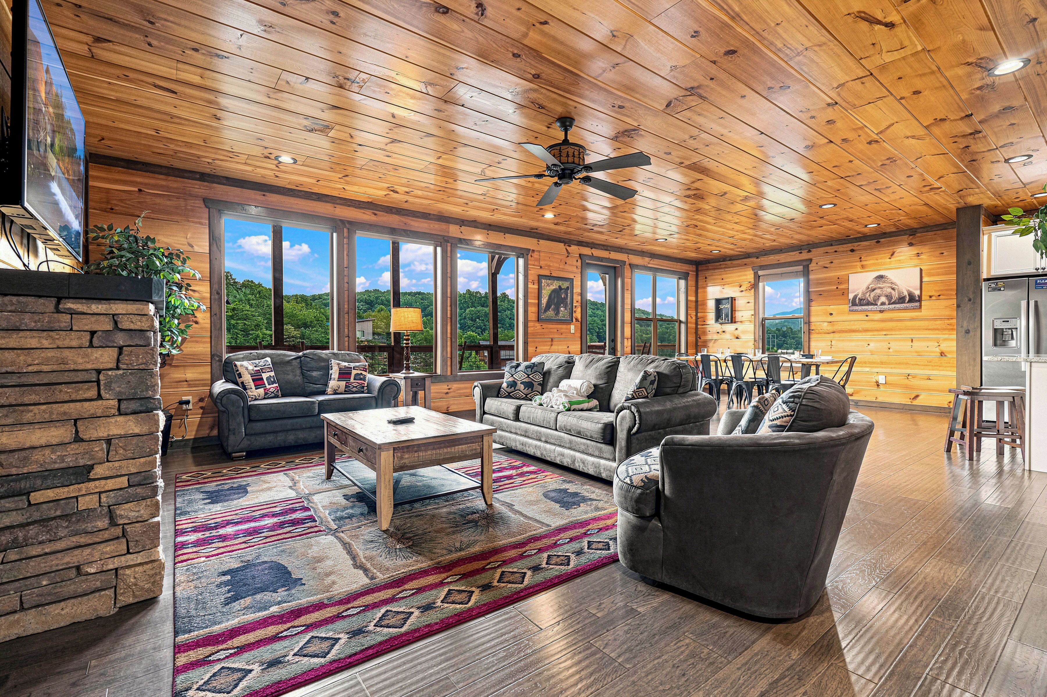 A spacious living room with a stone fireplace, large windows, and a ceiling fan.