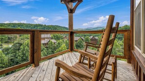 A wooden deck with a wooden chair, a metal railing, and a view of a green landscape with houses.