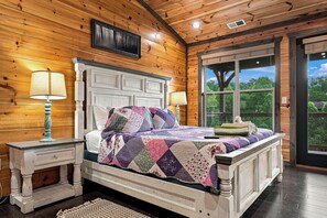 Quarto com painéis de madeira, uma cama, duas mesas de cabeceira e uma janela com vista para árvores.