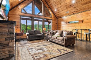 A spacious living room with a high ceiling, large windows, a stone fireplace, and a cozy seating area.