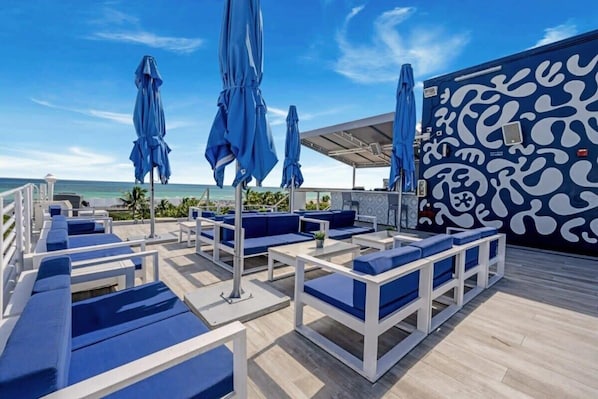 A rooftop lounge with blue cushioned seating and umbrellas, overlooking the ocean.