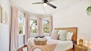 A bedroom with a bed, a ceiling fan, a window with curtains, and a bedside table with a lamp. - The Charleston Suites: The Emerson Suite (Charleston)