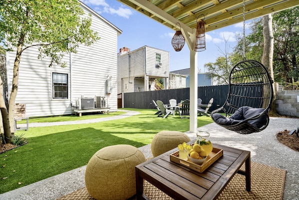 Ein Garten mit Holztisch, zwei Poufs, einem Hängesessel und Grillplatz.