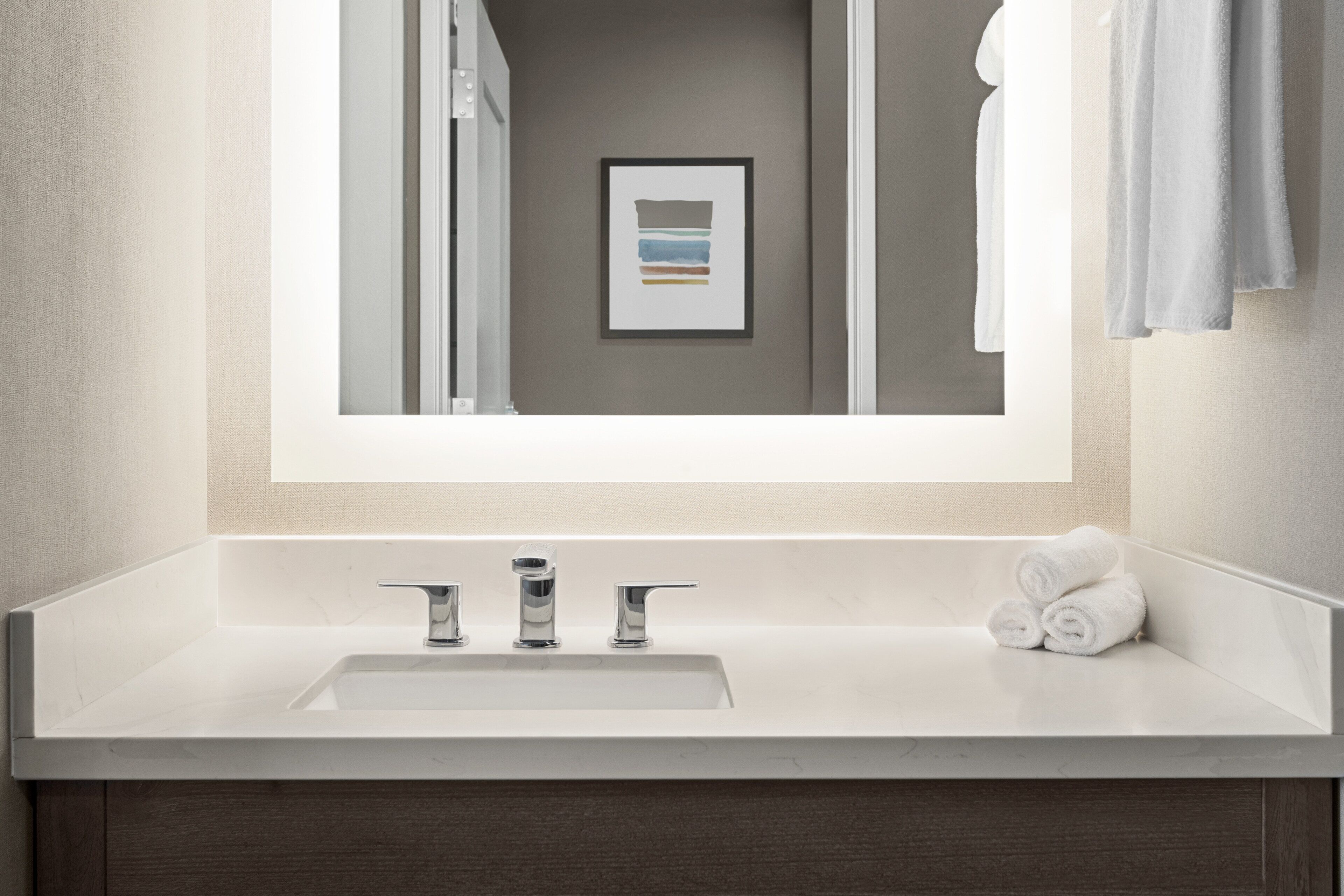 A modern bathroom with a white sink, three faucets, and a framed artwork on the wall.