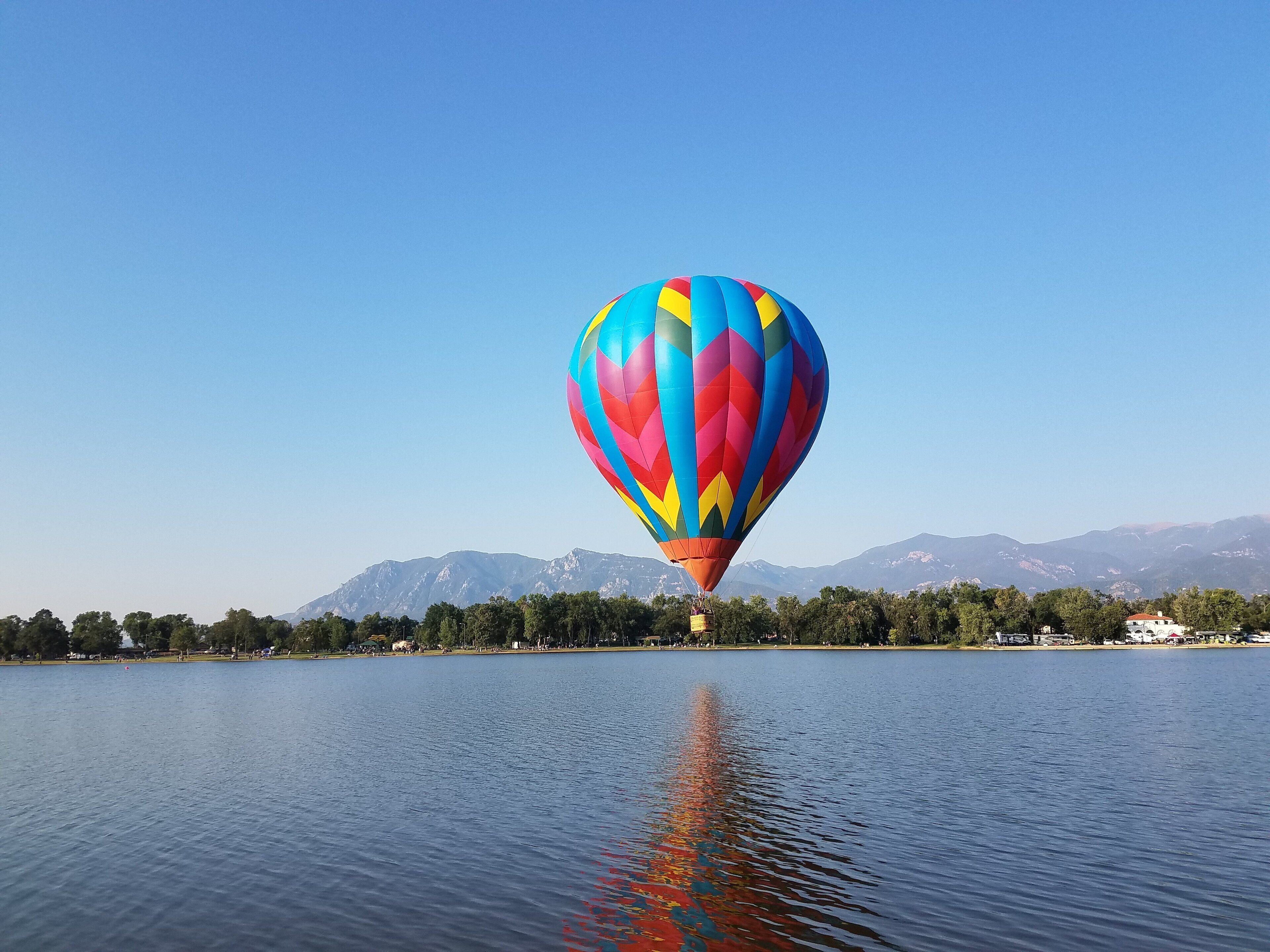 A hot air balloon over a calm lake with a clear sky and mountains in the background.