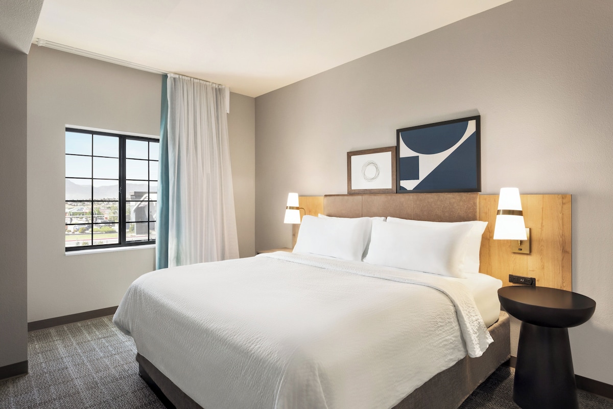 Quarto de hotel com uma cama grande, janela com cortinas, luminária fixada na parede e quadro emoldurado sobre a cama.