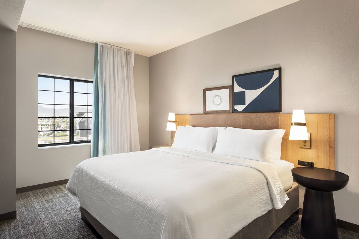 Una habitación de hotel con una cama grande, una ventana con cortinas, una lámpara de pared y un cuadro enmarcado sobre la cama.