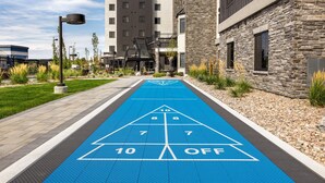 Uma quadra de shuffleboard azul com os números 10, 8, 7 e a inscrição 'OFF' visíveis, rodeada por edifícios e um lampião.