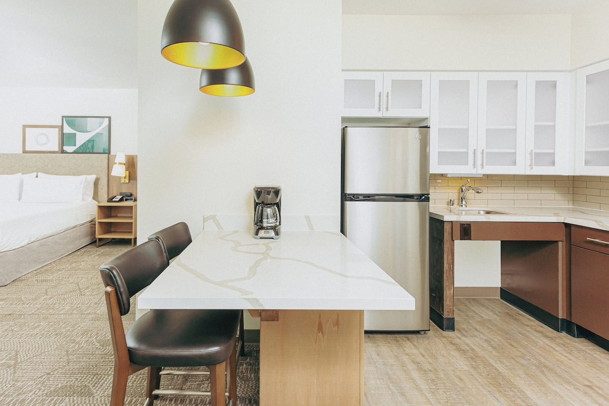Una habitación de hotel compacta con kitchenette, una cama con ropa de cama blanca, cafetera y una lámpara colgante moderna.