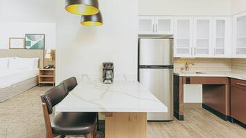 Habitación de hotel compacta con kitchenette, cama con ropa de cama blanca, mesa de comedor con sillas y una lámpara colgante moderna.