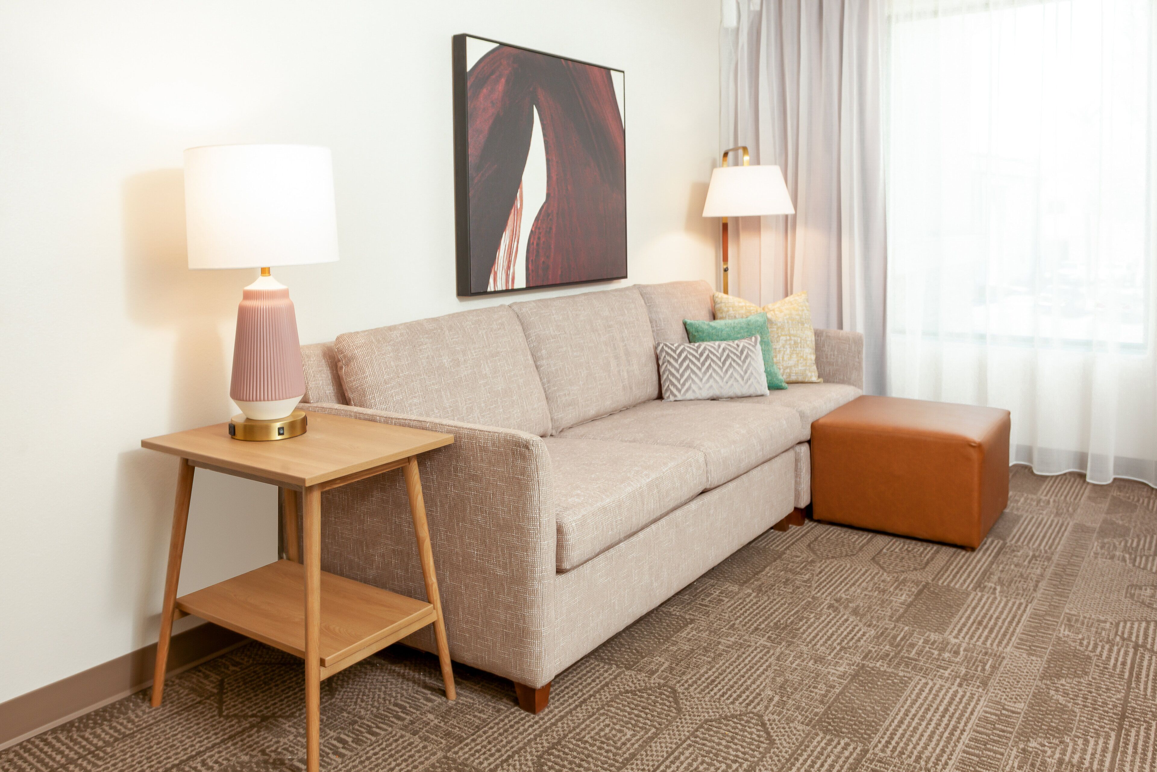 Habitación de hotel con un sofá color beige, una mesita de noche de madera con una lámpara, un reposapiés marrón, una alfombra estampada y un cuadro abstracto grande en la pared.