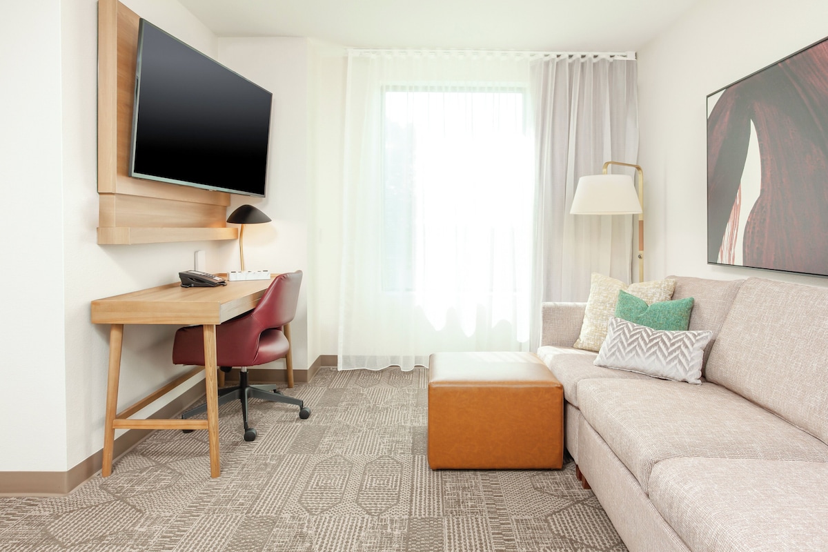 Habitación de hotel con un escritorio de madera, una silla de oficina roja, un sofá color beige, un reposapiés marrón, un televisor de pantalla plana grande montado en la pared, una lámpara de pie y una ventana con cortinas traslúcidas.