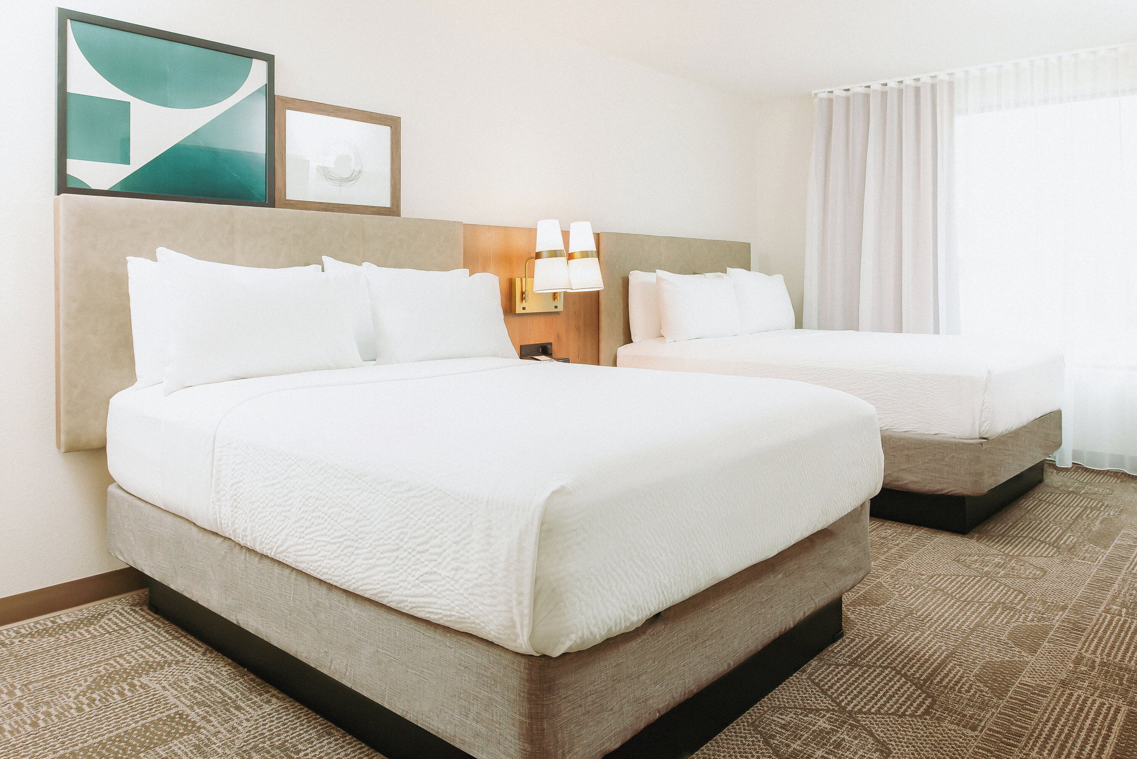 A hotel room with two beds, a headboard, a nightstand with a lamp, a framed picture, and a window with curtains.